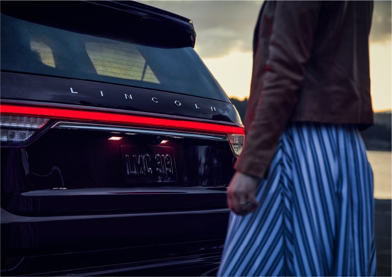 A person is shown near the rear of a 2023 Lincoln Aviator® SUV as the Lincoln Embrace illuminates the rear lights | Crossroads Lincoln of Southern Pines in Southern Pines NC